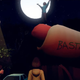 A man stands with his arms outstretched while standing on top of a giant hot dog with "bastard" engraved on its bun.
